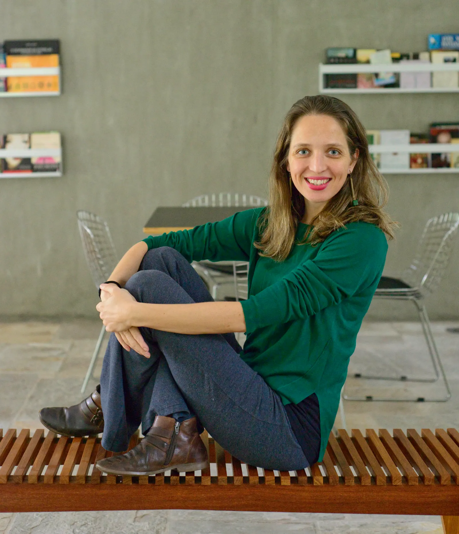 Mirella Duarte sentada em um banco de madeira, diante de uma parede de concreto com prateleiras de livros